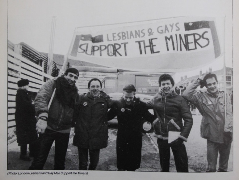 A black and white picture of five people in winter coats holding up and standing under a banner that reads, Lesbians & Gays Support the Miners.