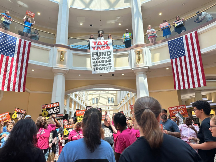 A crowd of people stand in a large hall at the Pennsylvania state capitol holding up signs. Up on a balcony, more people are seen holding signs and three people hold a large banner that reads, Tax Yass, Fund Education, Housing, Health Care, Transit 