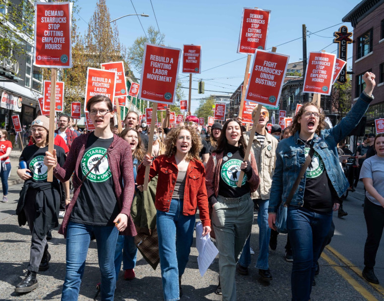 A crowd of people march down the middle of a street holding up red signs with pro-union messages. The people in the foreground are wearing black  t-shirts featuring the Starbucks Workers Union logo.